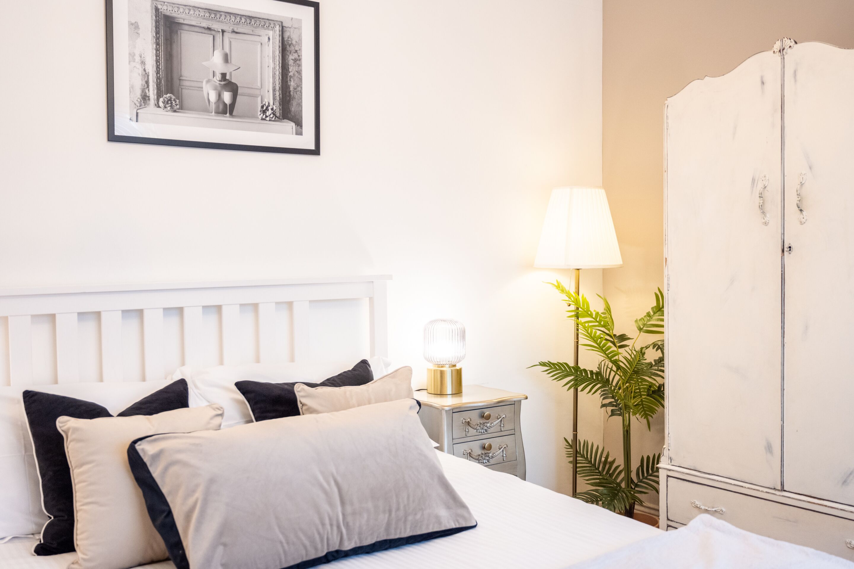 A neatly made bed with a headboard, a bedside table with a lamp, a wardrobe, and a framed picture on the wall.