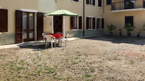 A building with a courtyard, featuring a table with chairs under a green umbrella, potted plants, and a balcony with a railing. - Casa Olinda (Lazise)