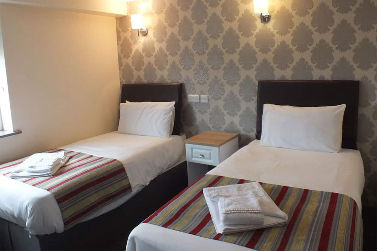 A hotel room with two single beds, each with a striped bedspread and white pillows. There are bedside tables with lamps and a small drawer. The walls have a patterned wallpaper.