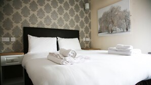 A neatly made bed with white linens and rolled towels, a nightstand with a drawer, and a painting of trees on the wall. - The Fazeley Inn (Tamworth)