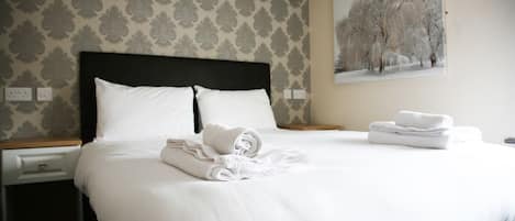 A neatly made bed with white linens and rolled towels, a nightstand with a drawer, and a painting of trees on the wall.