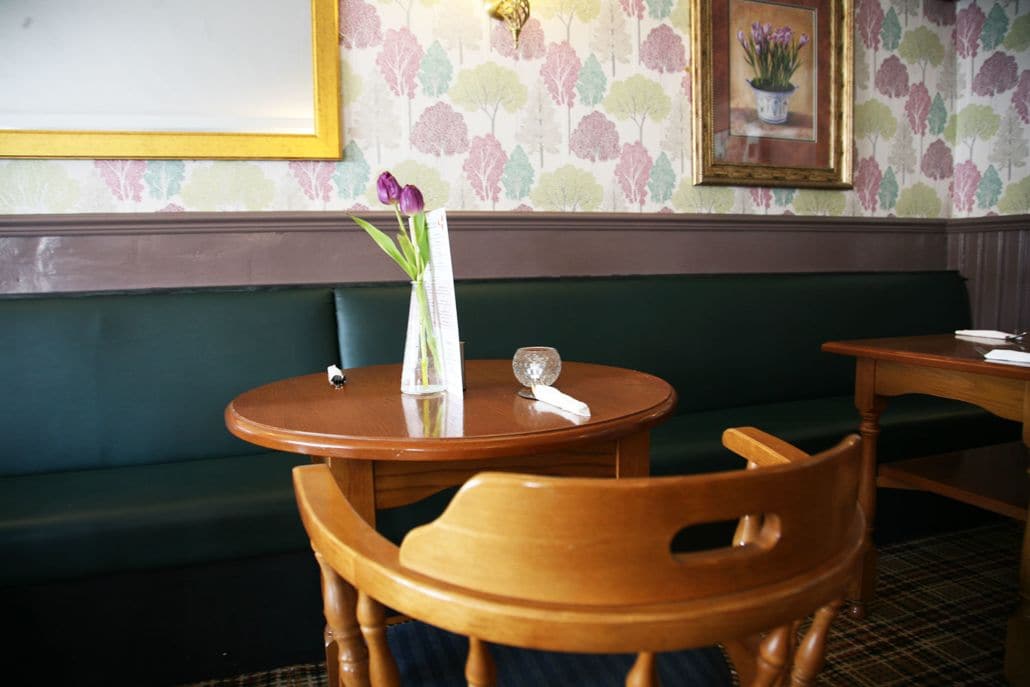 Una habitación con un banco tapizado en verde, una mesa redonda de madera con un florero y un cuadro en la pared.