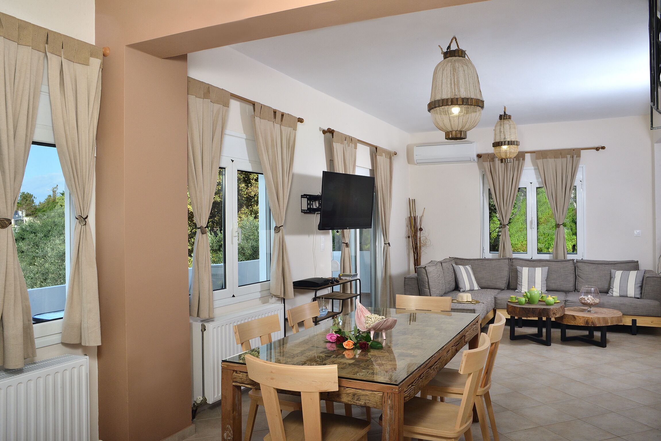 Un comedor con una mesa de vidrio, sillas de madera y un sofá. Hay un televisor sobre un mueble bajo y cortinas en las ventanas.