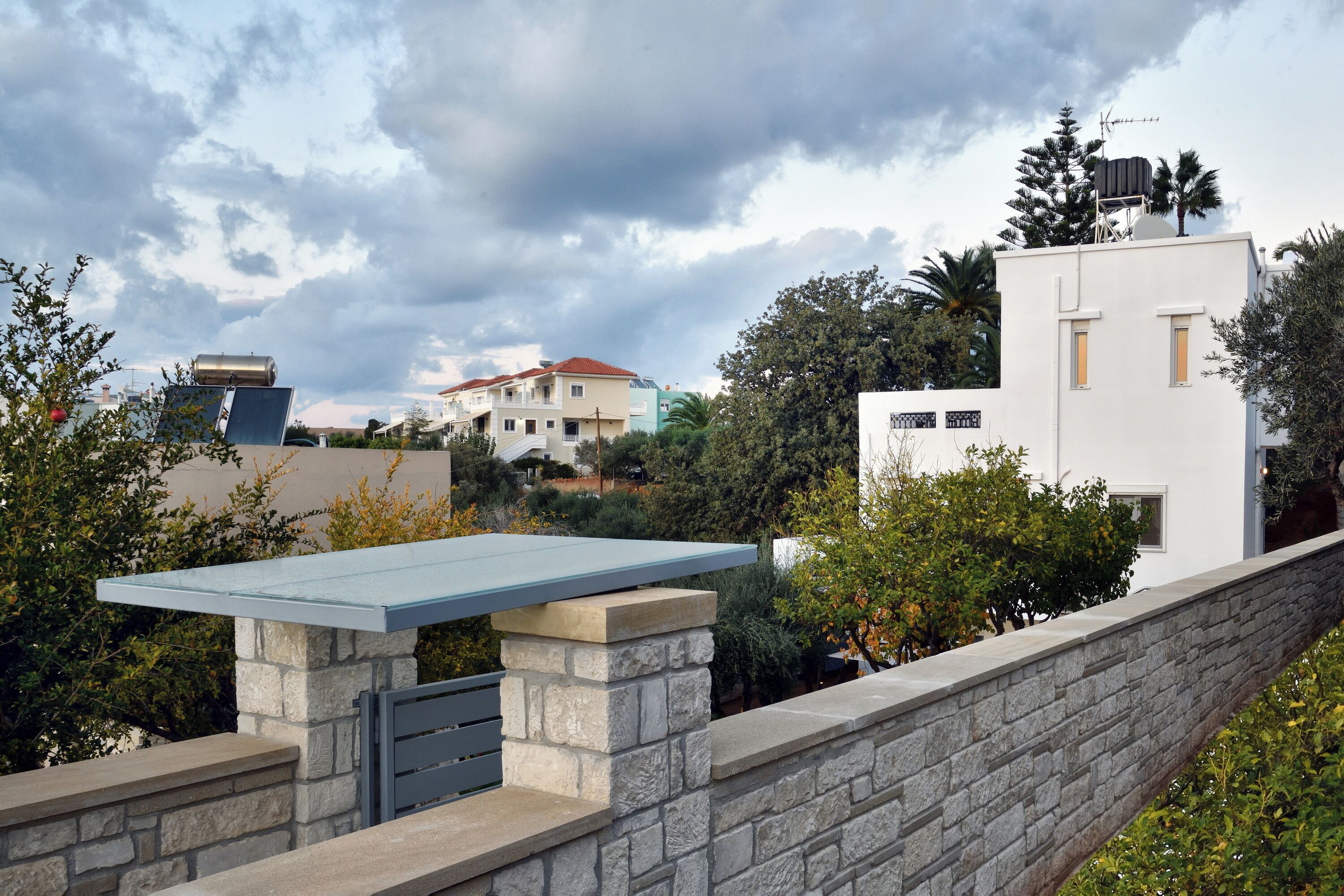 Zona d'estar exterior moderna amb pared de pedra i taula de vidre, envoltada de vegetació i edificis residencials.