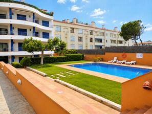 A swimming pool with lounge chairs, a well-maintained lawn, and a multi-story building with balconies. - Apartament Vip Ancla first line of the sea (Escala, L')