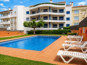 A swimming pool with lounge chairs, a tree, and a multi-story building with balconies.