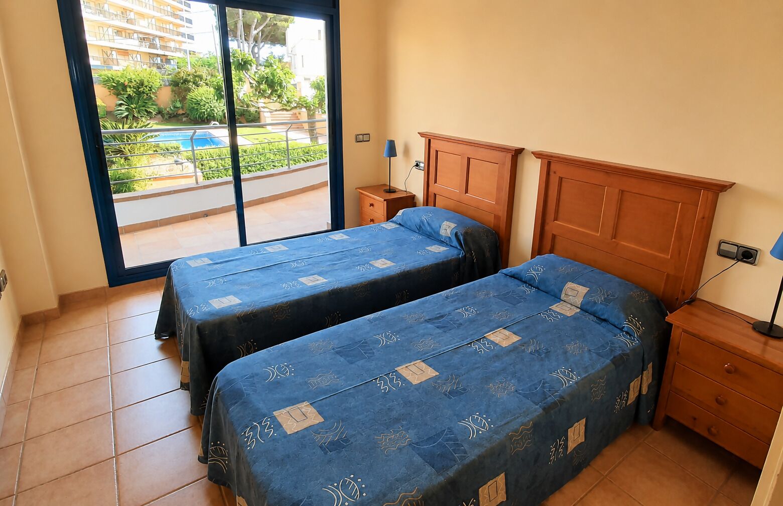 A bedroom with two beds, a wooden headboard, a nightstand, a lamp, and a view of a pool and greenery outside.