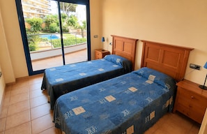 Ein Schlafzimmer mit zwei Betten, einem Holz-Kopfende, einem Nachttisch, einer Lampe und Blick auf einen Pool und Grünflächen.