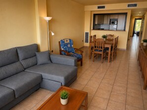 A living room with a sofa, a dining table with chairs, a lamp, and a kitchen area with cabinets and appliances.