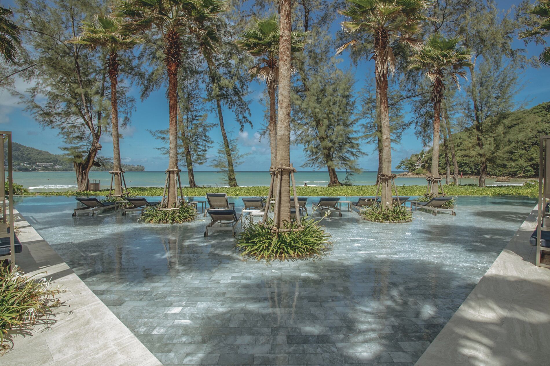 A poolside area with lounge chairs and palm trees, overlooking a beach and ocean.
