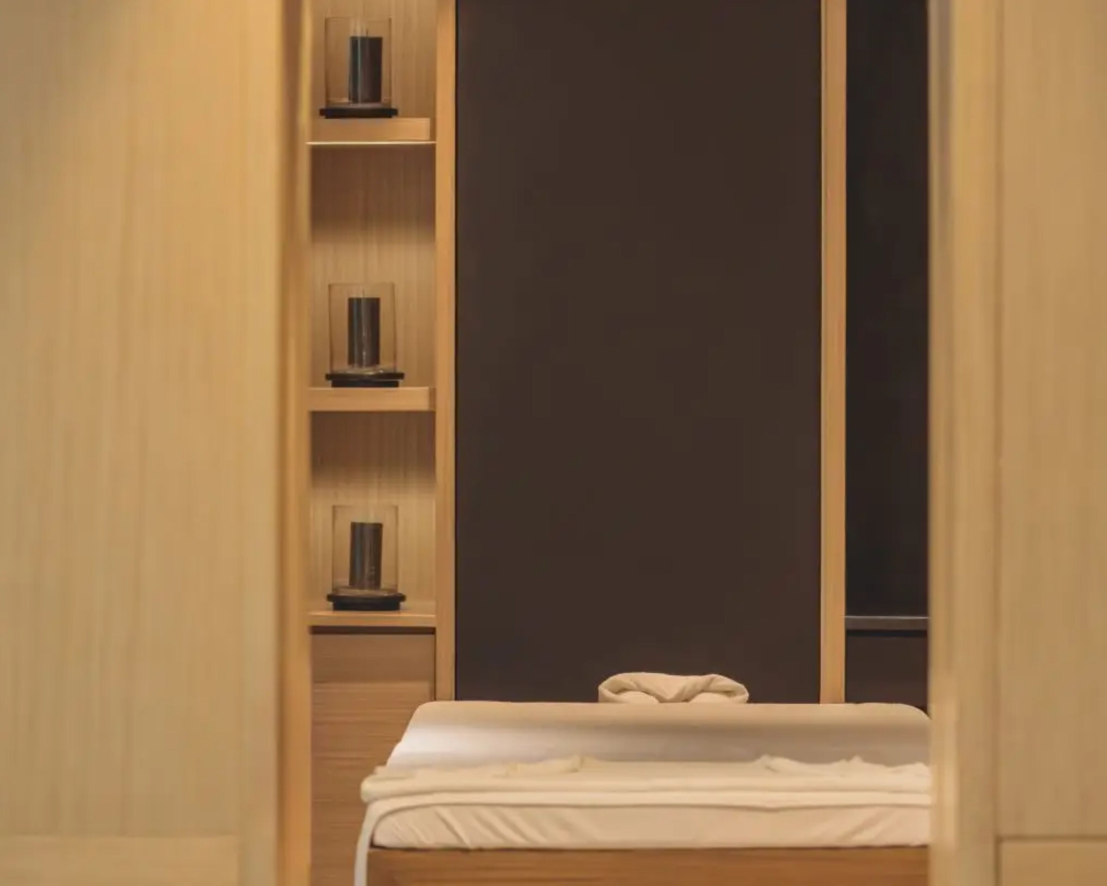 A hotel room with a bed, a wooden wardrobe with shelves, and a dark-colored door.