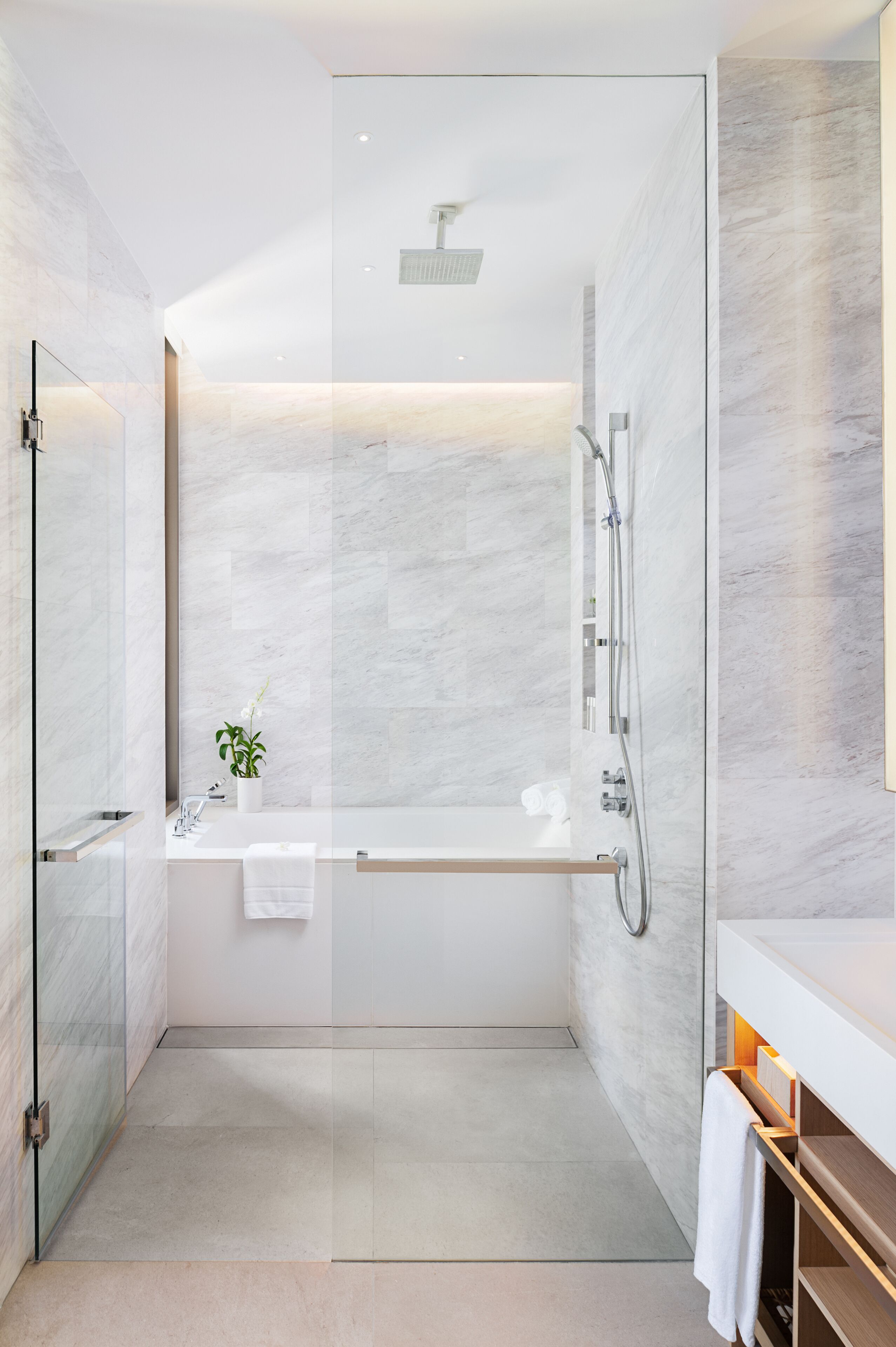 Un baño moderno con ducha envuelta en cristal, pared de mármol blanco y tocador de madera con toallero.