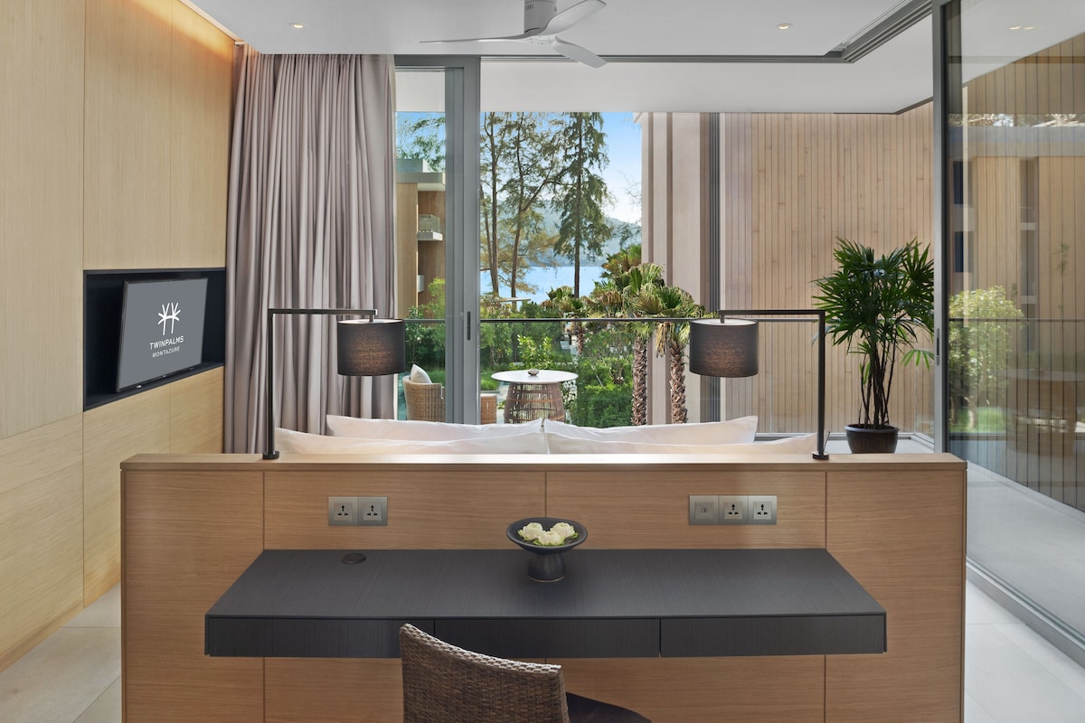 Una habitación de hotel moderna con un escritorio de madera, un frutero, una planta en maceta y vistas a un balcón con mobiliario exterior y vegetación.
