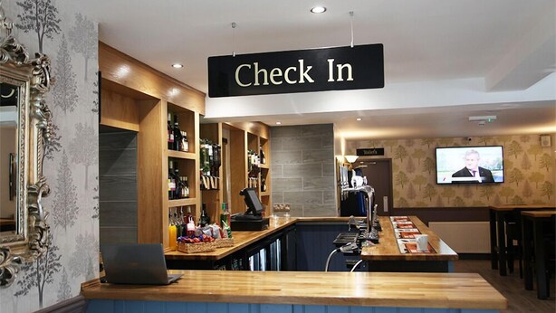 Un mostrador de recepción con un cartel que dice 'Check In', una computadora portátil y una zona de bar con botellas y vasos.