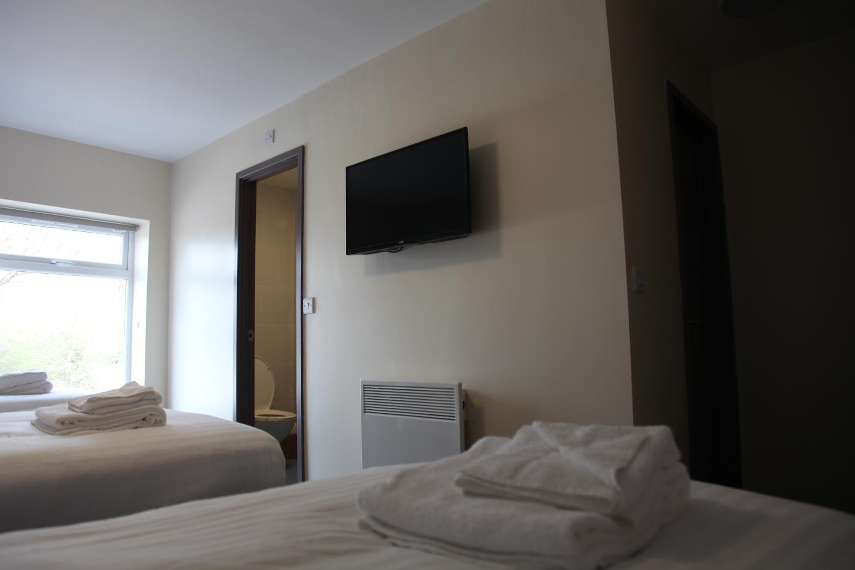 Habitación de hotel con dos camas, un televisor montado en la pared, una ventana y una puerta que da a un baño.