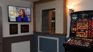 Una habitación con televisión mostrando un noticiero, una máquina tragamonedas, cuadros en la pared y un radiador.