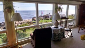 A room with large windows overlooking a rocky coastline, featuring a rocking chair, a table with a lamp, and potted plants. - Quintessential Maine waterfront! (Biddeford)