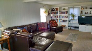 A living room with a leather sofa, a television, a bookshelf, and a window with plants. - Quintessential Maine waterfront! (Biddeford)