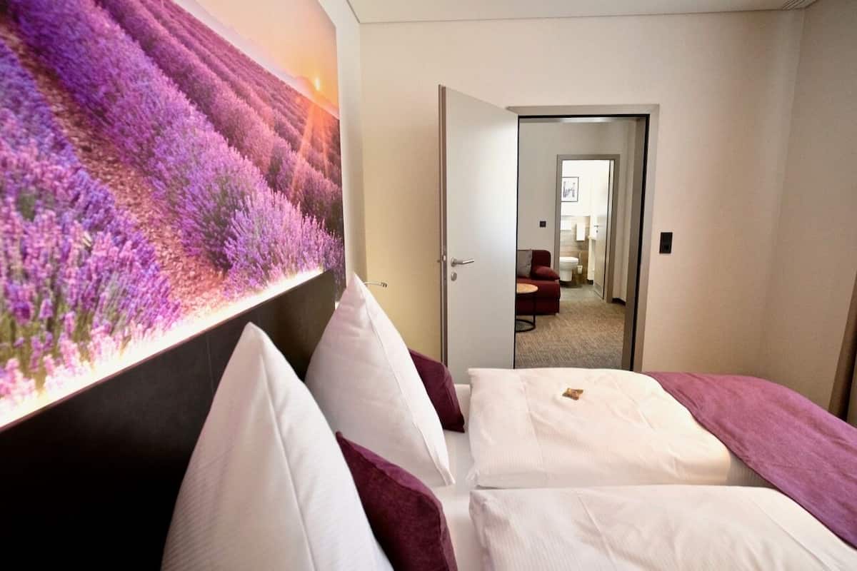 A hotel room with a large bed, a headboard with a floral mural, and a view into the hallway.
