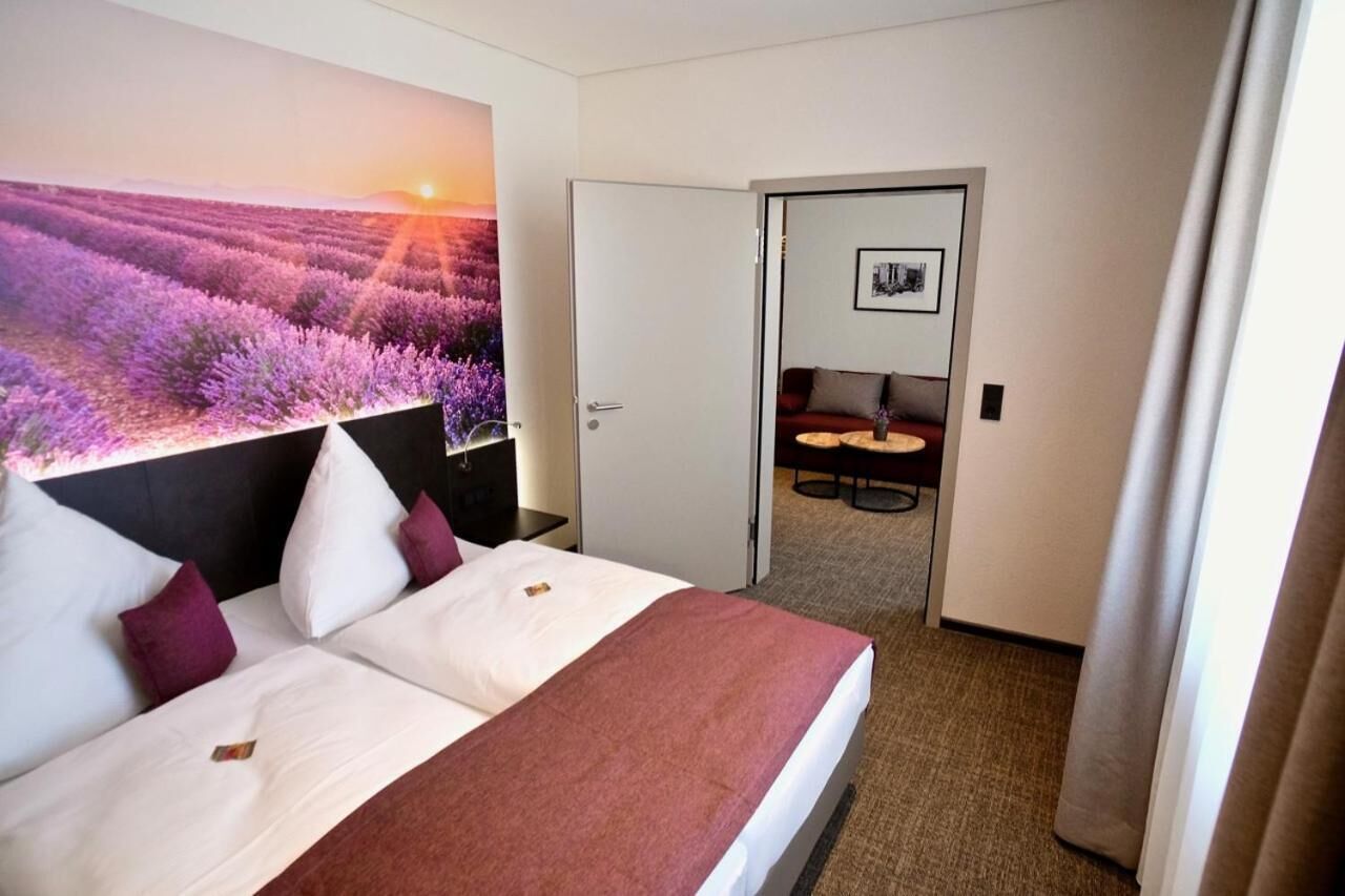 Une chambre d’hôtel avec un lit, une table de chevet, un bureau et une chaise. Il y a un tableau au mur et une porte qui donne sur une autre pièce.