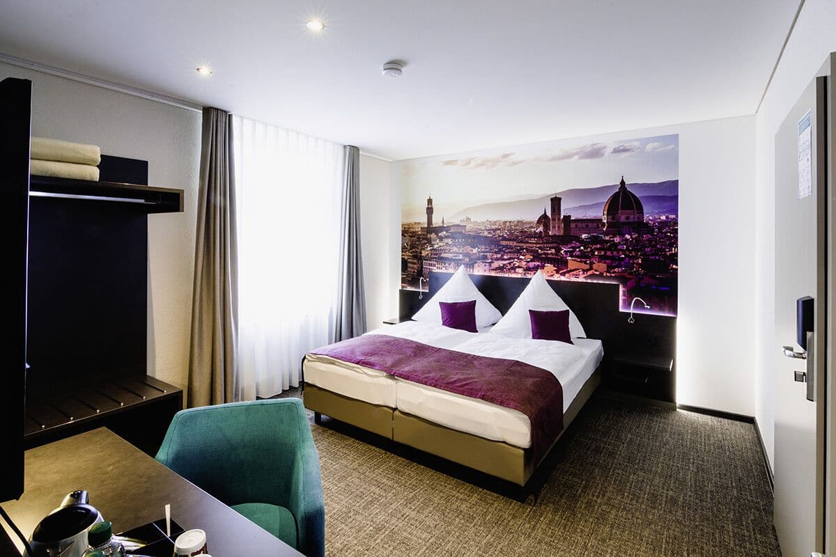 A hotel room with a large bed, a desk with a chair, a wardrobe, and a cityscape view.