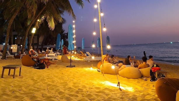 Ein Strand mit Palmen, Liegestühlen und Sonnenschirmen, beleuchtet durch Lichterketten.