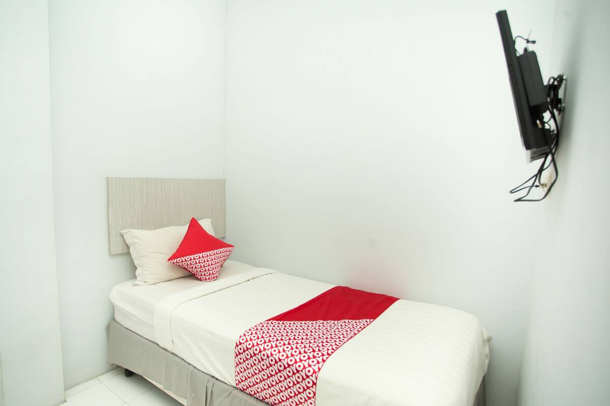 Une petite chambre avec un lit simple, une couvre-lit blanche et rouge, une tête de lit grise et un téléviseur fixé au mur.