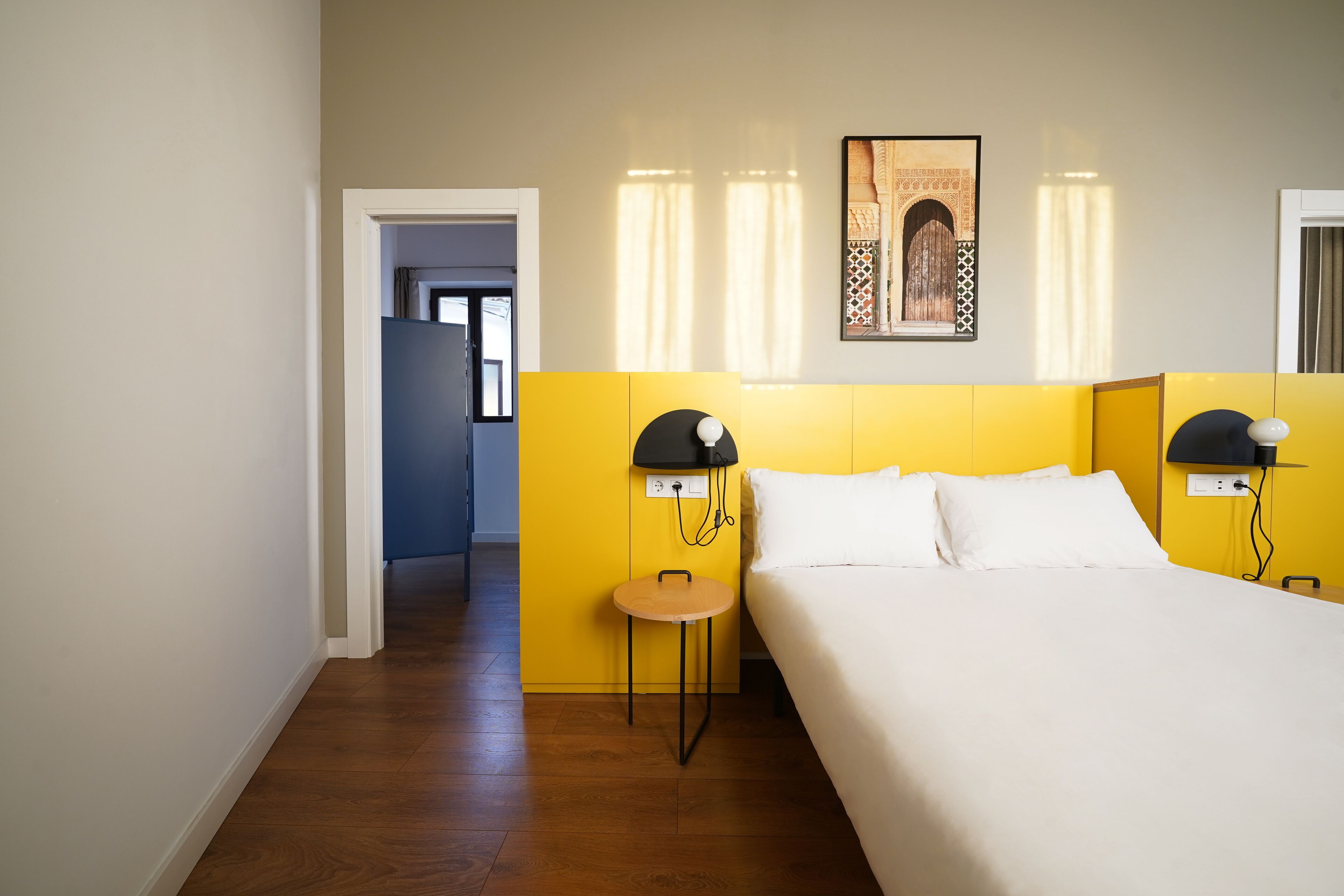 Hotelkamer met een bed, een klein rond tafeltje en twee bedlampen. Er hangt een ingelijste foto aan de muur en een deur die leidt naar een andere kamer.