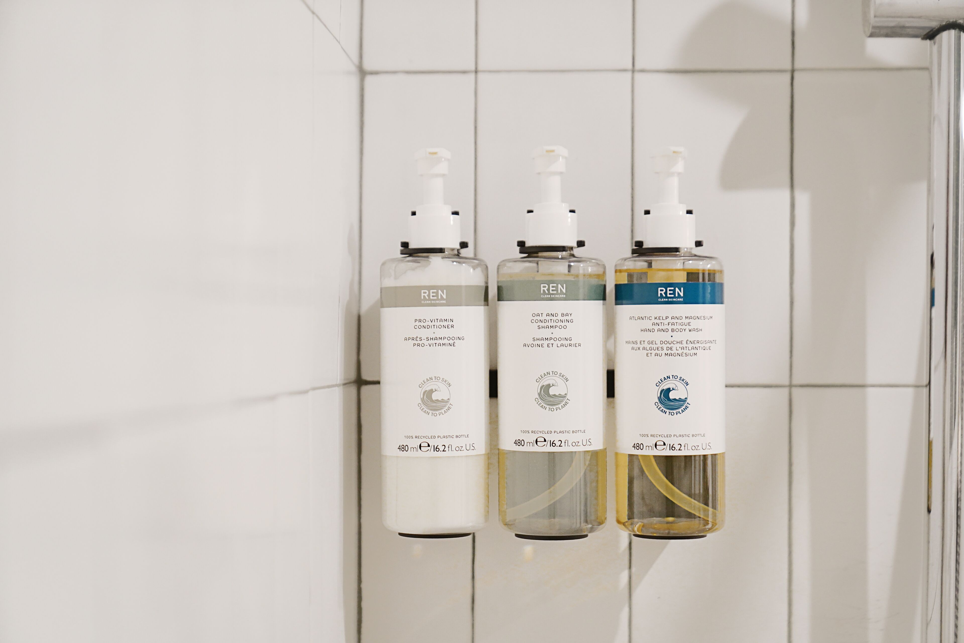 Tres botellas de productos REN sobre una pared de azulejos blancos.