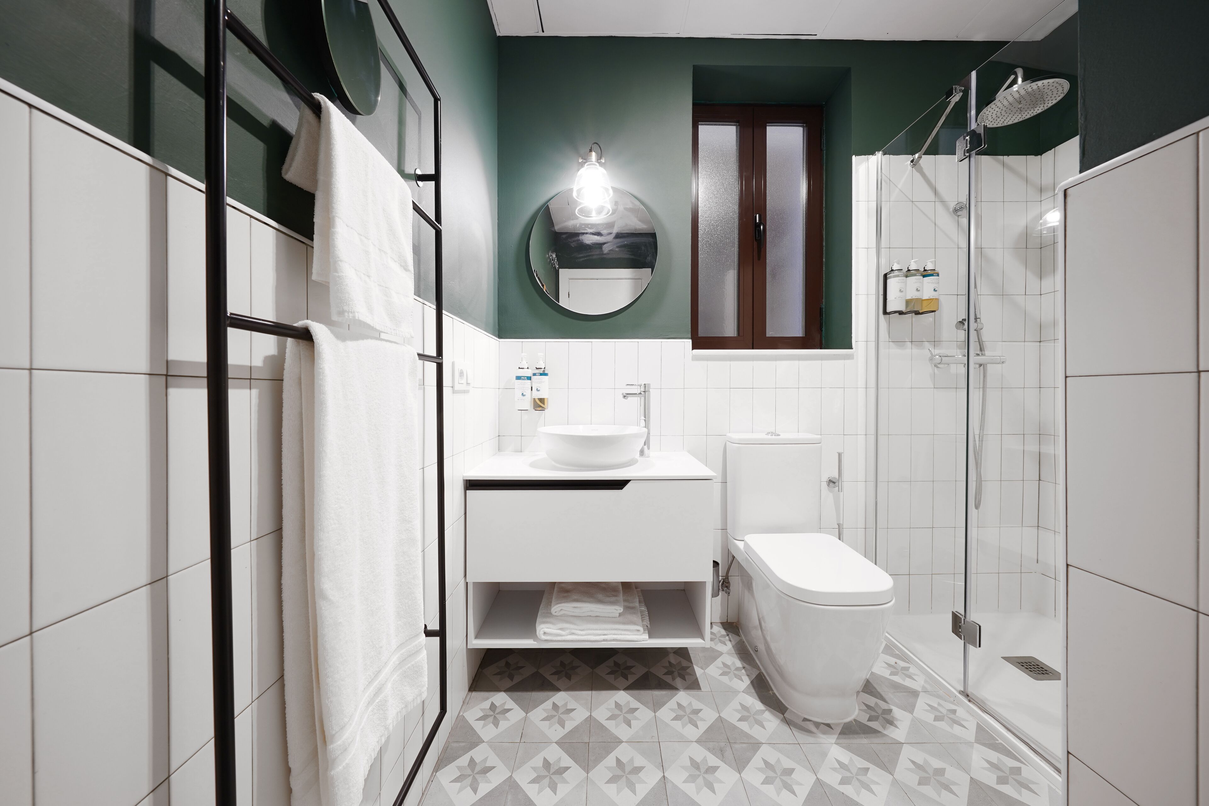 A modern bathroom with a white sink, toilet, and shower. It features a round mirror, a towel rack, and a tiled floor with a geometric pattern.
