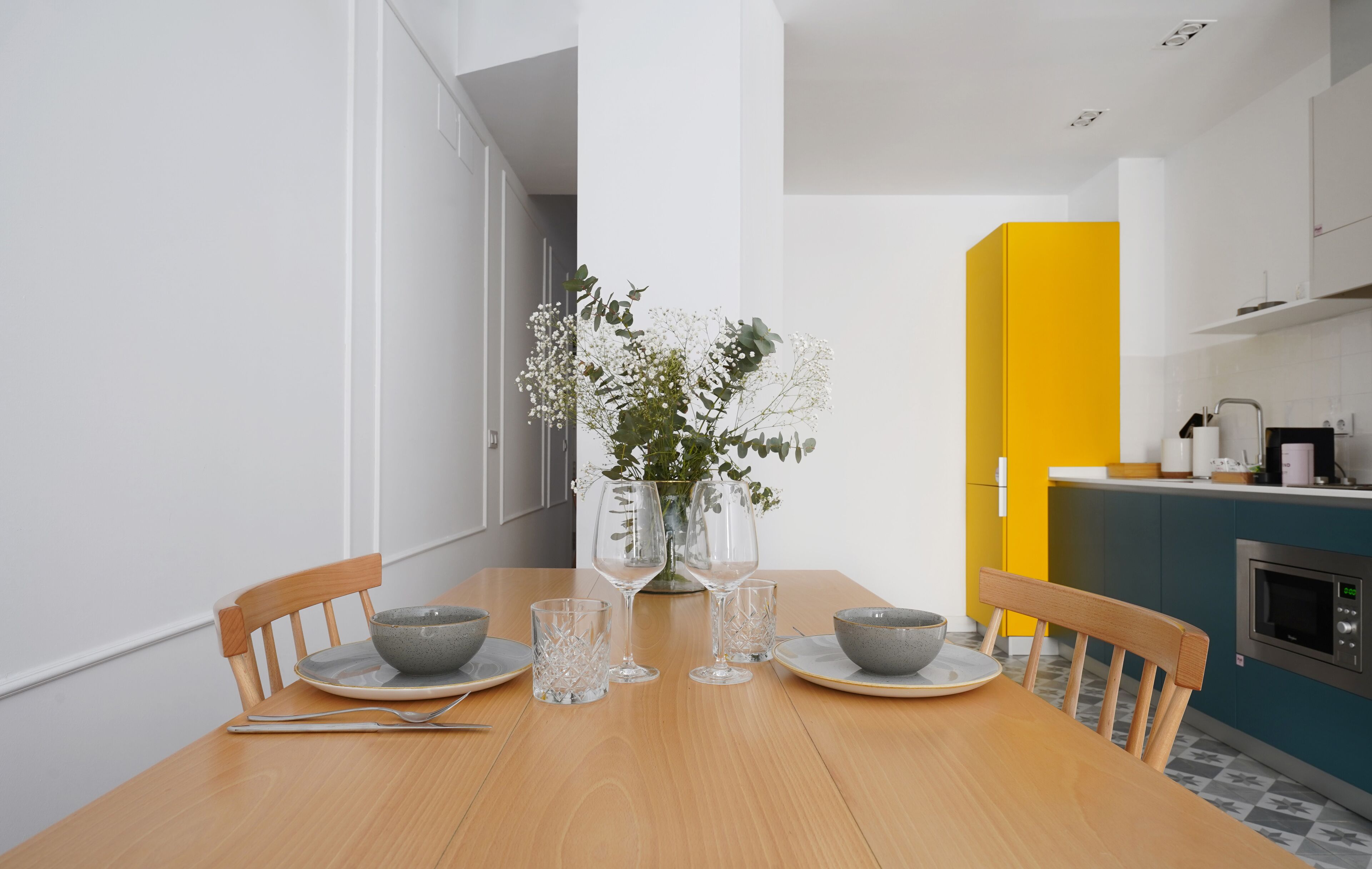 A dining area with a wooden table set for two, a vase with flowers, and a kitchen in the background.