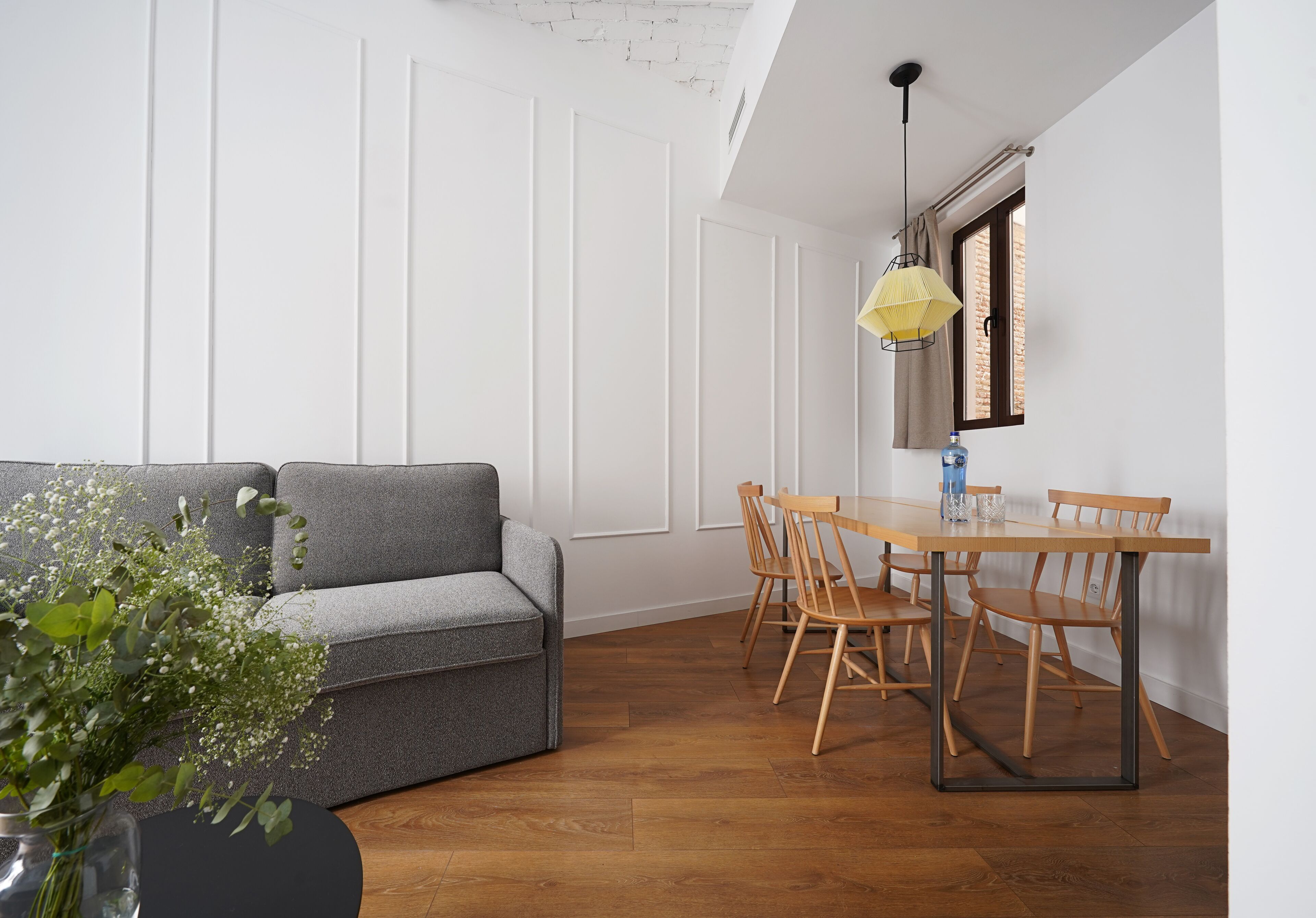 Una sala moderna con un sofá gris, una mesa de comedor de madera y sillas. Hay un jarrón con flores en el suelo.