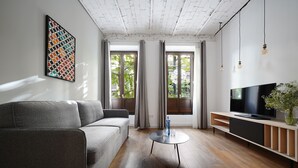 A modern living room with a grey sofa, a round coffee table, a TV on a wooden stand, and large windows with curtains. - Numa Granada Vega (Granada)