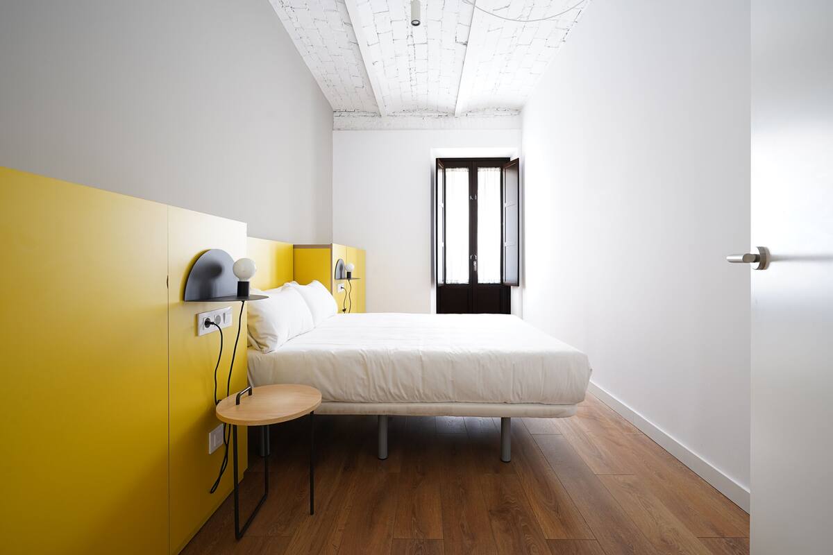 A modern bedroom with a bed, bedside table, lamp, and a view of the outside through a window.