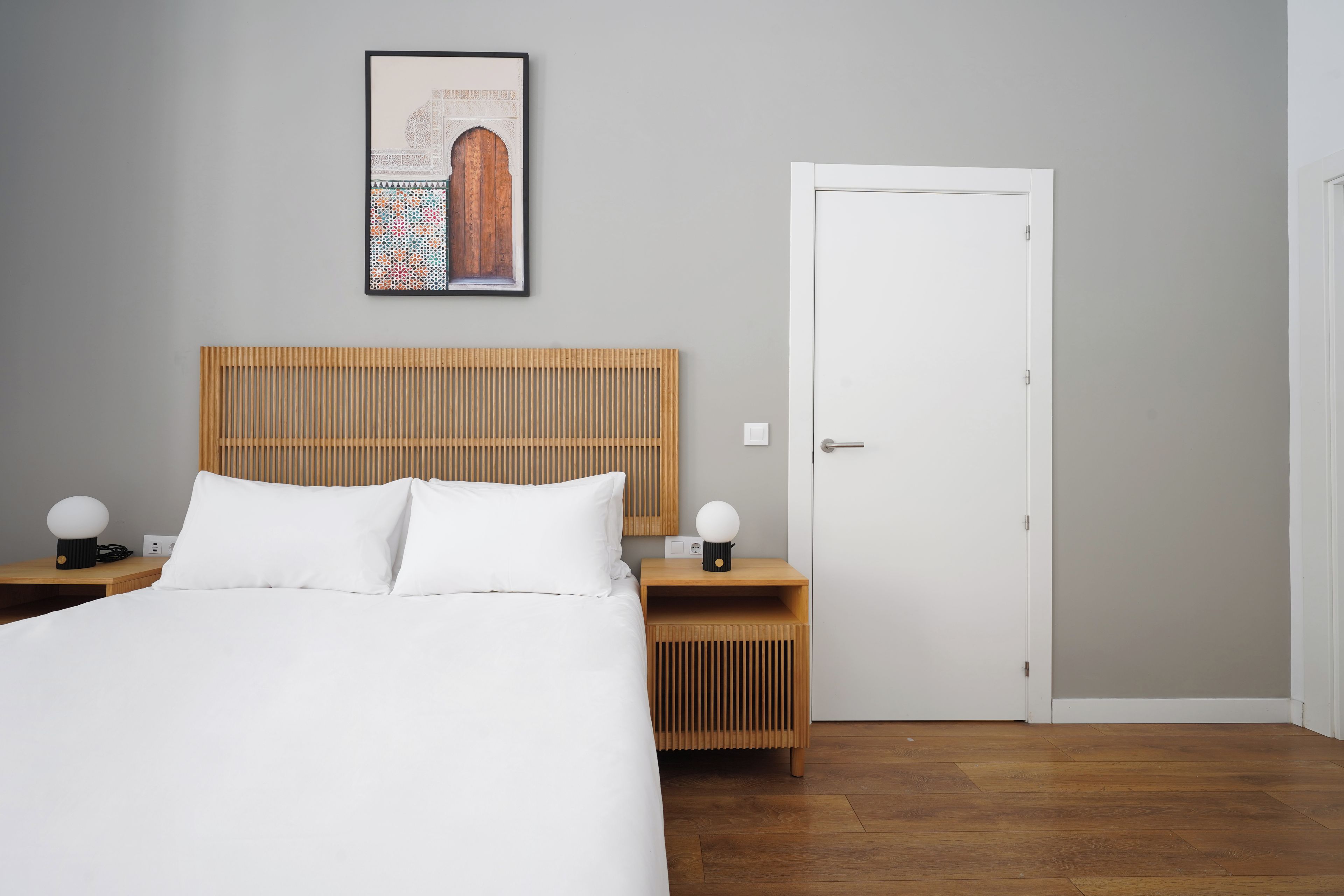 A modern bedroom with a wooden headboard, a white bed, two bedside tables with lamps, and a door.