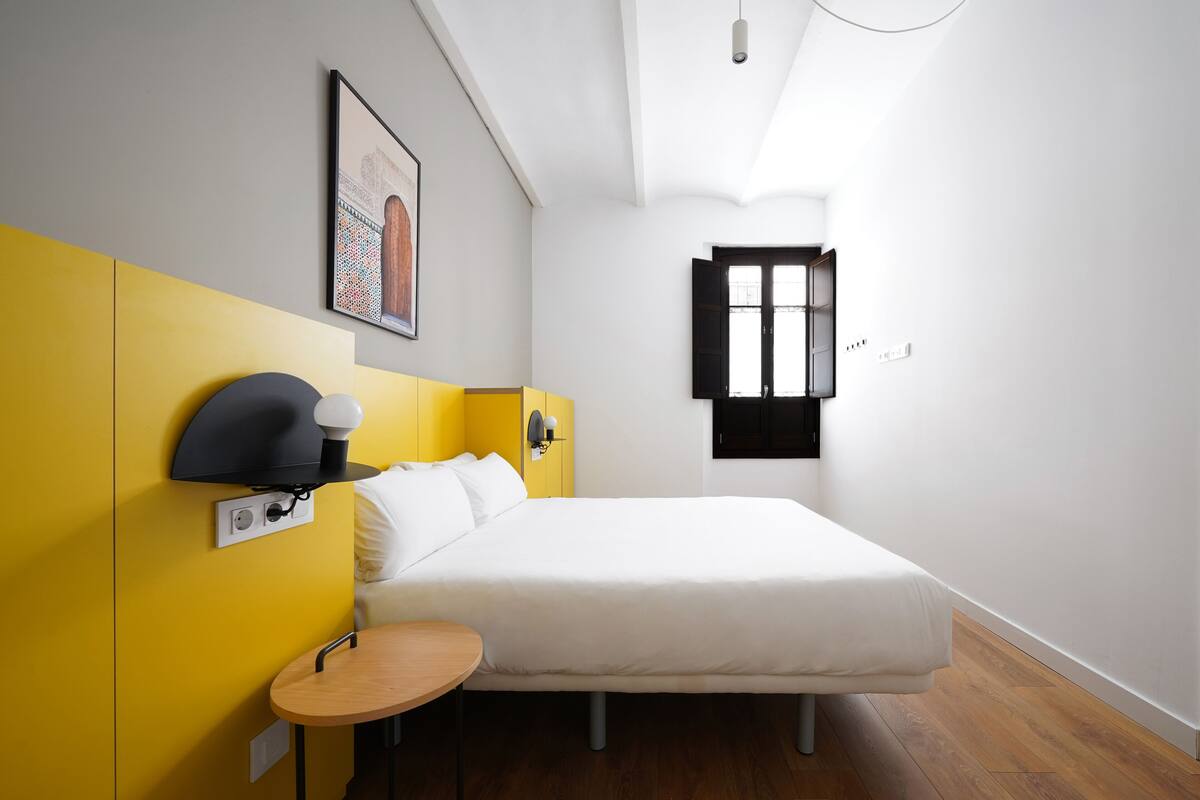 A bedroom with a bed, a bedside table, a wall-mounted lamp, a window, and a picture on the wall.