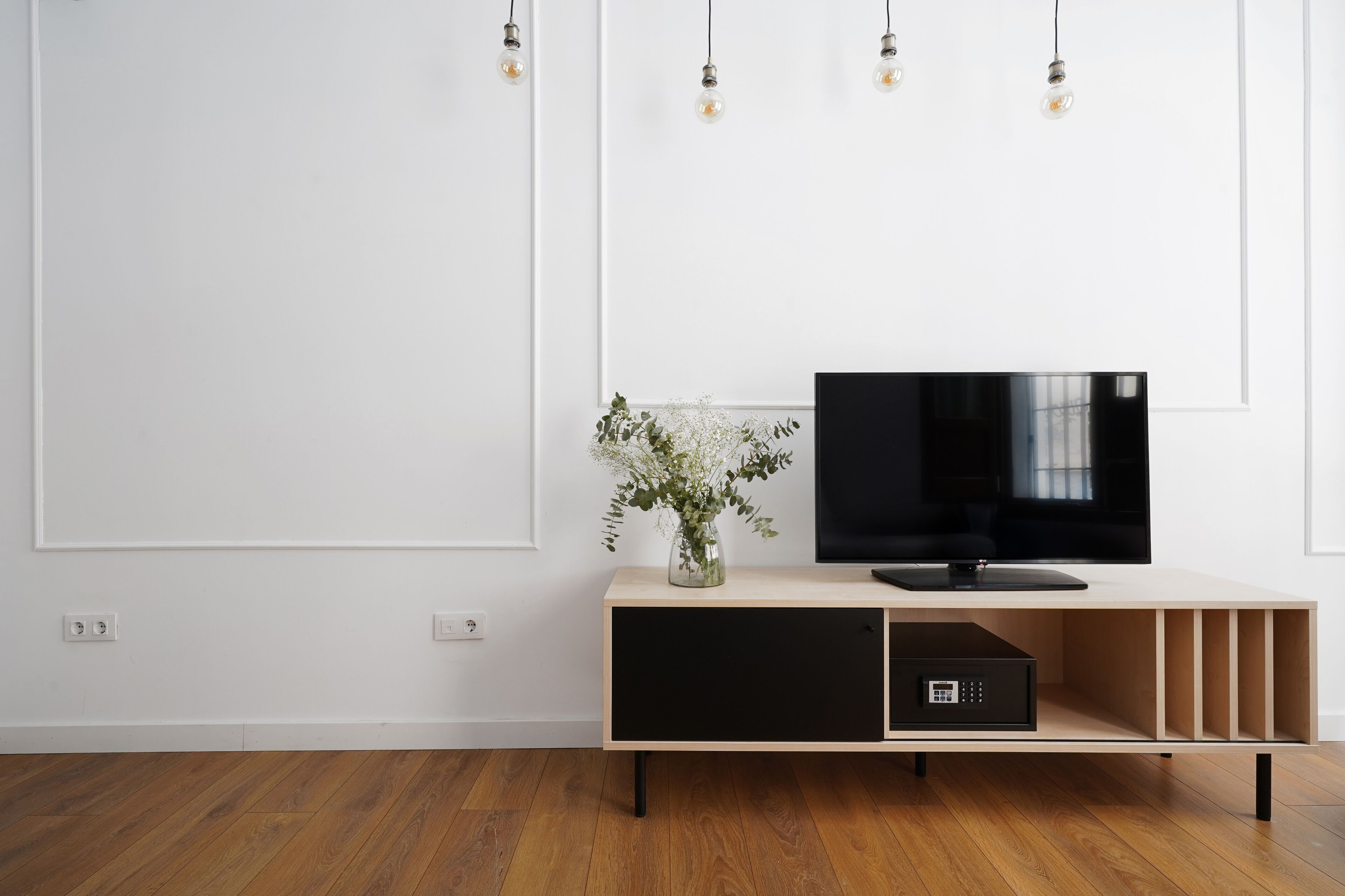 A modern living room with a wooden floor, a TV on a light-colored console, and a vase with flowers.