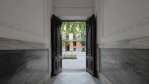 A hallway with a marble floor and dark wooden doors leading to an outdoor area with a view of buildings and greenery. - Numa Granada Vega (Granada)