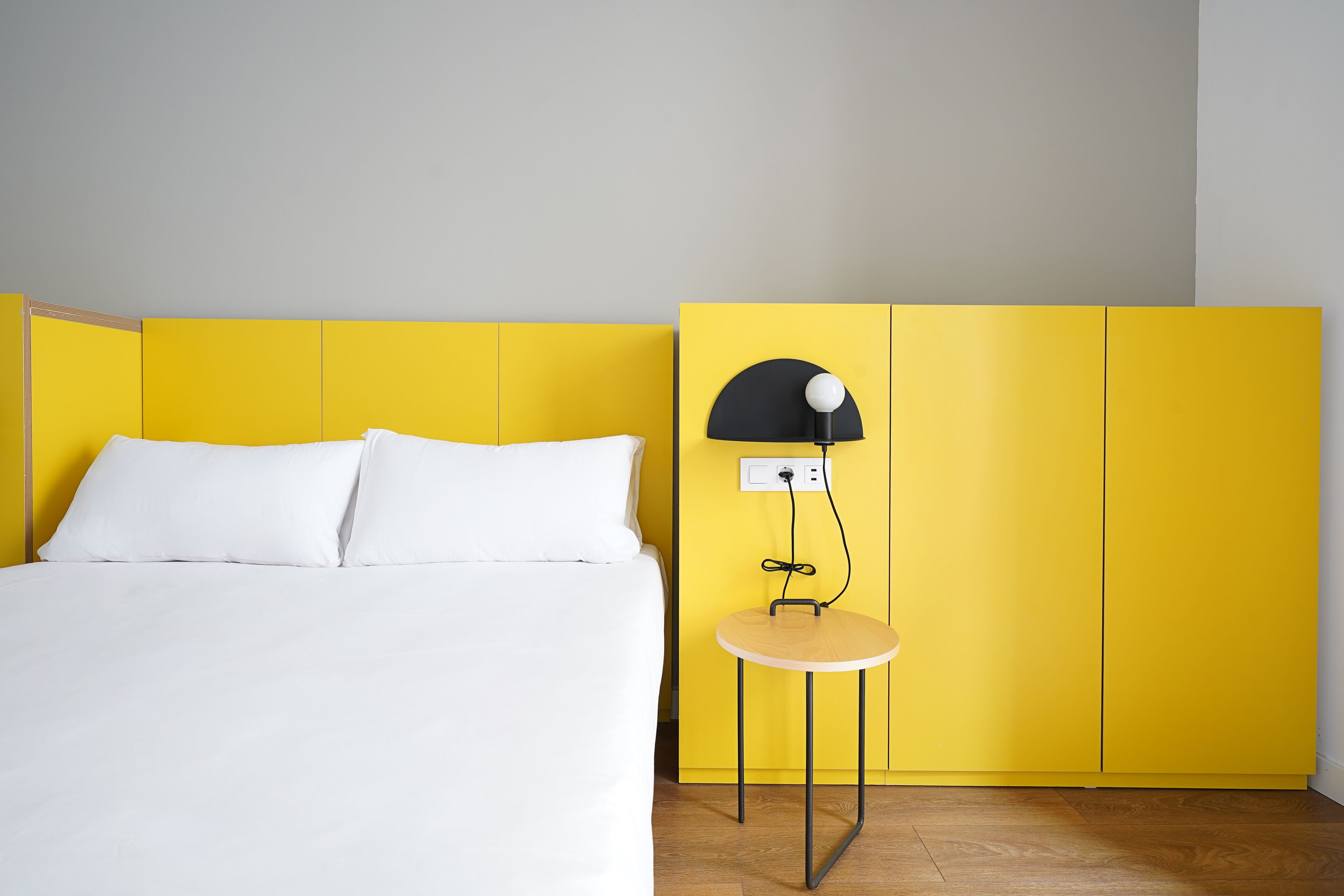 A bed with white linens, a yellow wall, and a small table with a lamp.