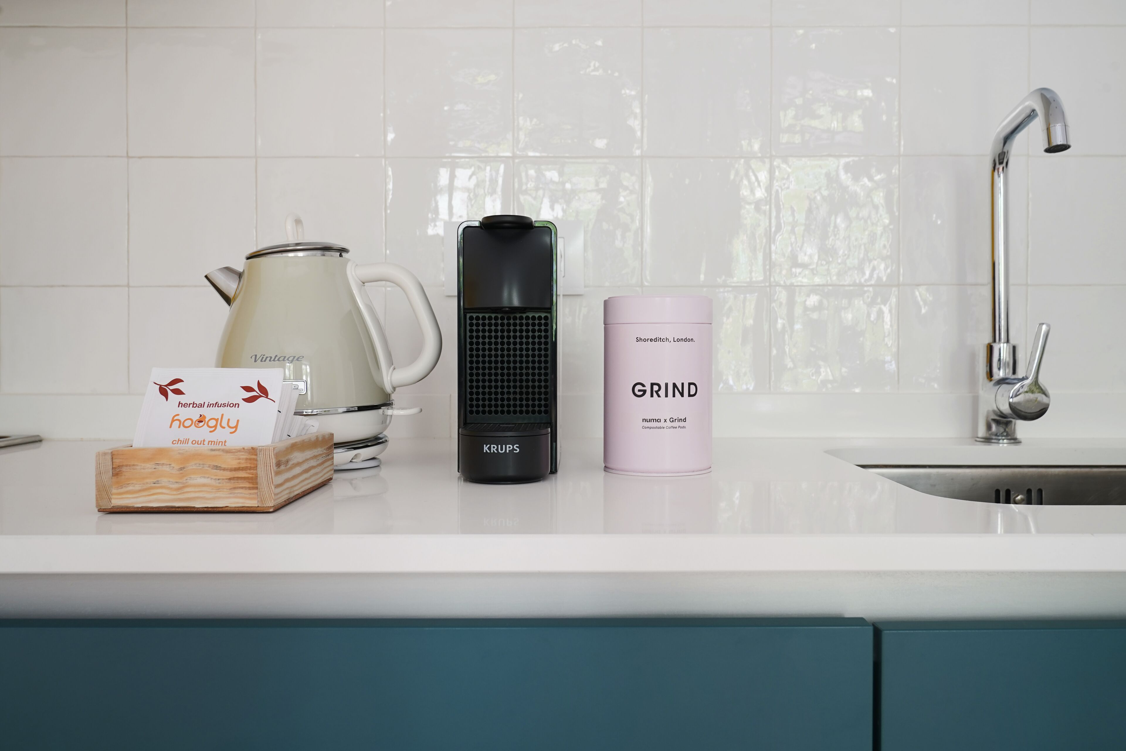 Una encimera de cocina con una tetera, una cafetera, un recipiente con la etiqueta 'SHERIDITCH, LONDON, GRIND' y una caja con la inscripción 'infusión herbal hoagly chill out menta'.