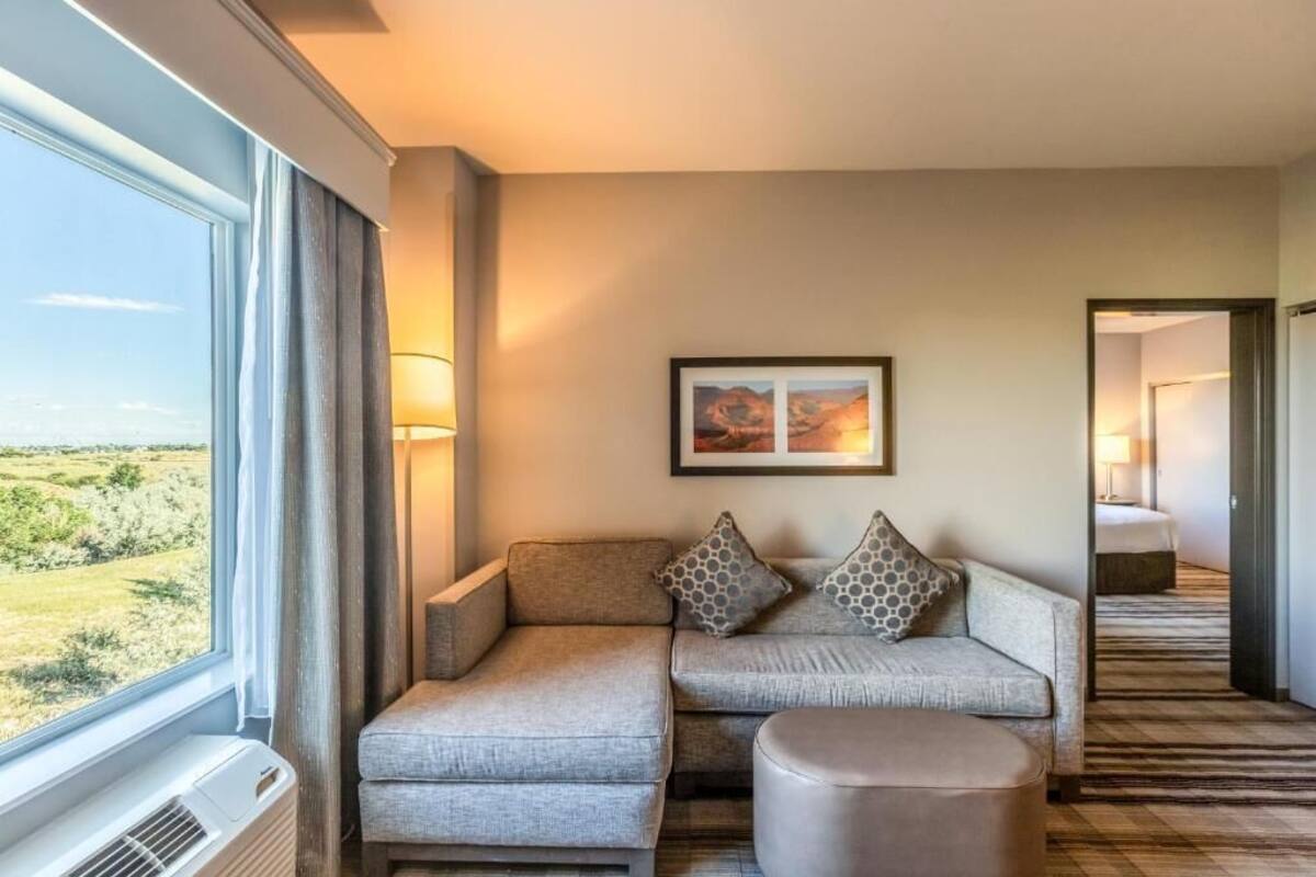 Een hotelkamer met een groot raam, een bank, een voetenbankje en een lijst met een afbeelding aan de muur.