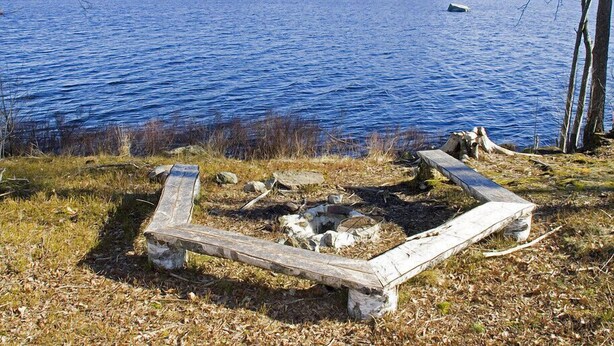 A lakeside scene with a wooden fire pit surrounded by rocks and a bench. - Holiday Home Sundslätt (Urshult)