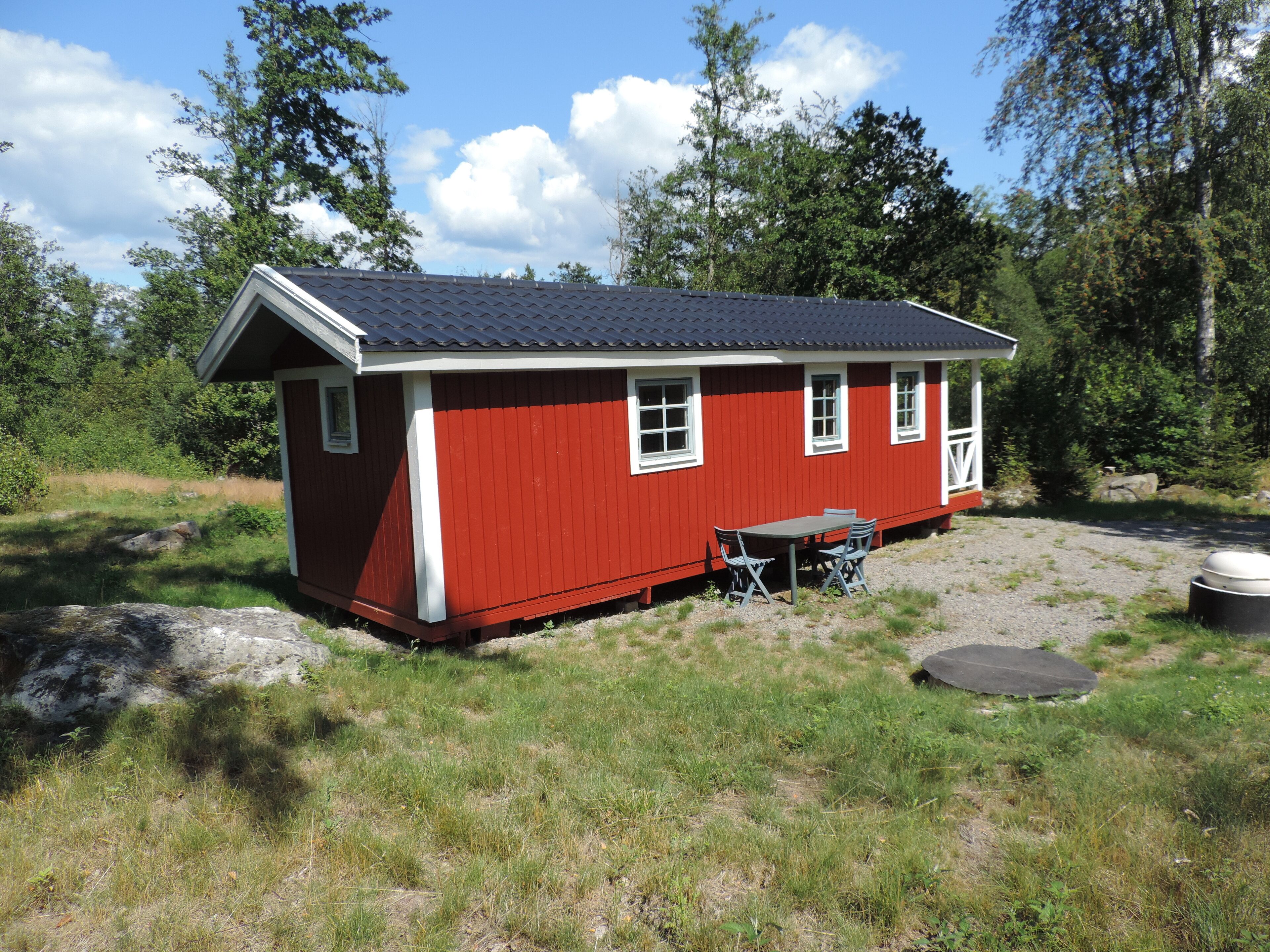 A small, red wooden cabin with a dark roof, a table with chairs, and a large round object nearby.