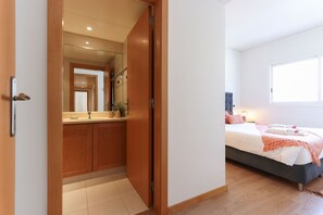 A bedroom with a bed, a bedside table, a window, and a bathroom with a mirror and sink. - Estrela Design by Homing (Lisbon)