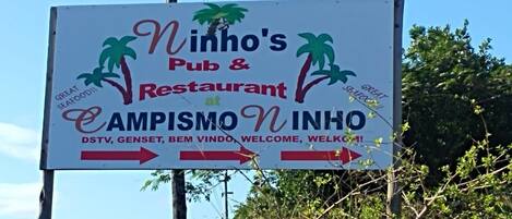 A sign for Ninho's Pub & Restaurant at CAMPISMO INHÓ, with directional arrows and welcoming text.
