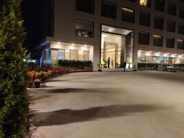 Entrada moderna de un hotel por la noche con fachada iluminada, zona exterior cubierta con plantas en macetas y estacionamiento.