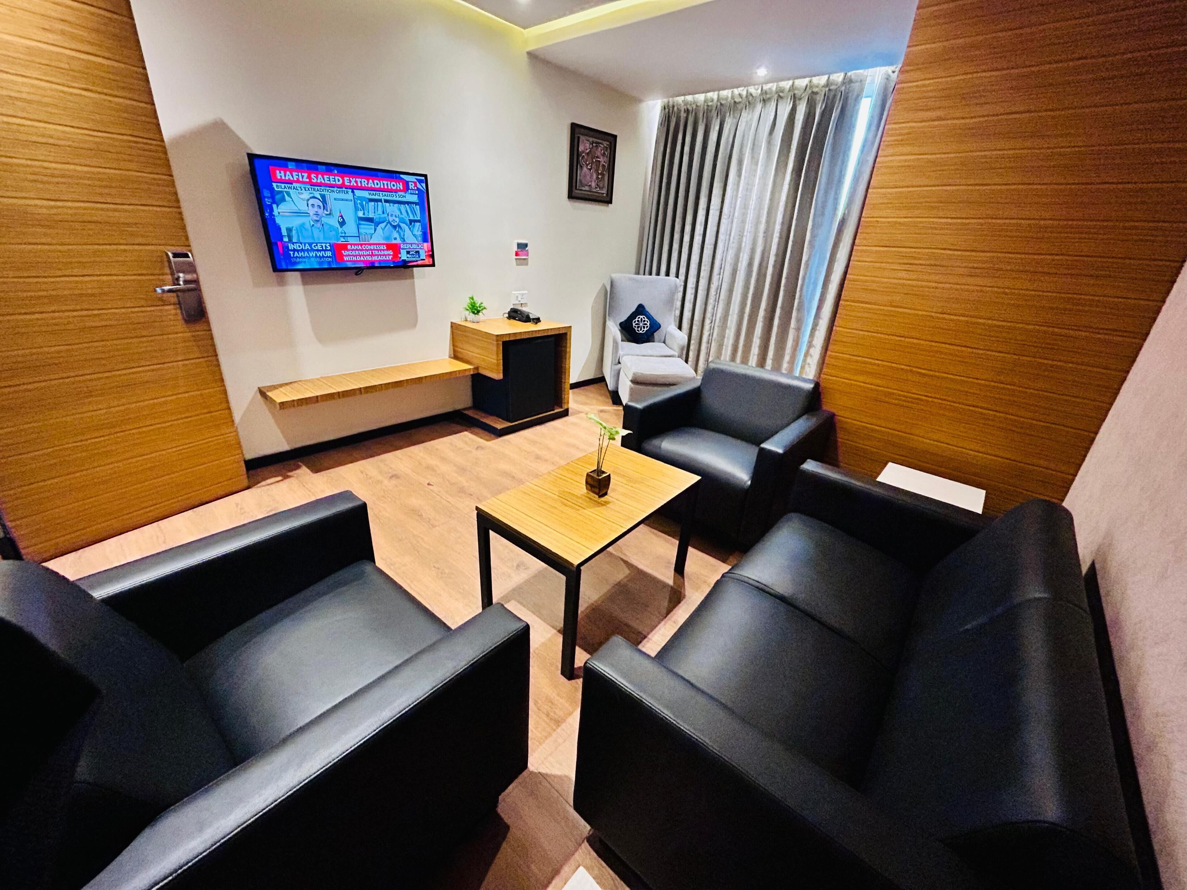 A modern living room with black leather sofas, a wooden coffee table, a TV mounted on the wall, and a small side table with a plant.