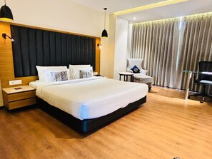 A hotel room with a large bed, a nightstand, a chair, a small table, and a window with curtains. - Novanta Jamshedpur (Adityapur)