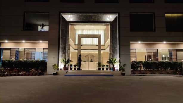 A modern building entrance with a glass facade, potted plants, and a person standing in the doorway. - Novanta Jamshedpur (Adityapur)