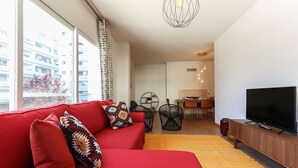 Un salon moderne avec un canapé rouge, des fauteuils noirs, un téléviseur et, au fond, un coin repas.